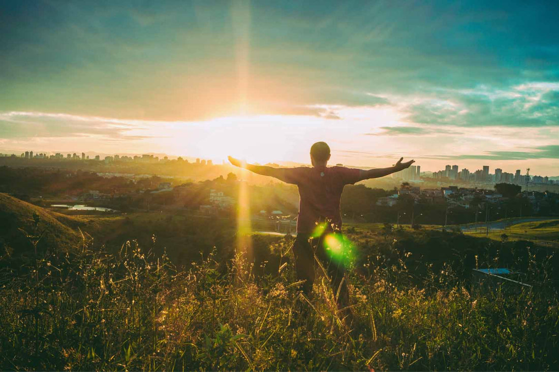 Mensch mit ausgebreiteten Armen auf einem Feld zum Sonnenuntergang blickend