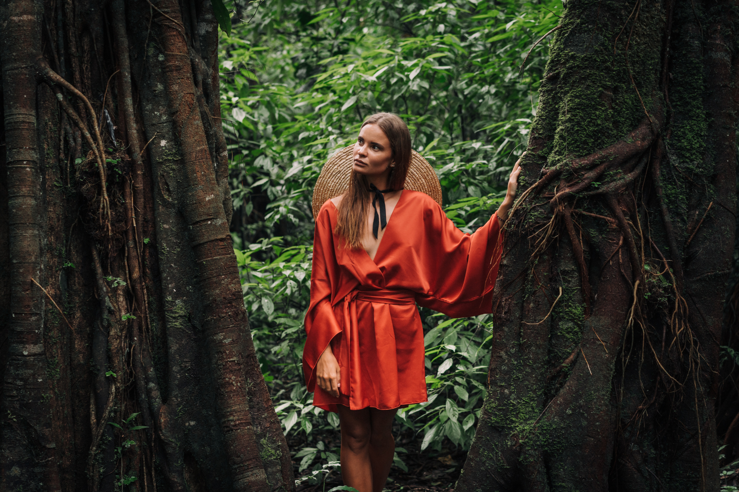 Frau in rotem Kimono aus Baumwollsatin im Dschungel steht zwischen zwei Bäumen