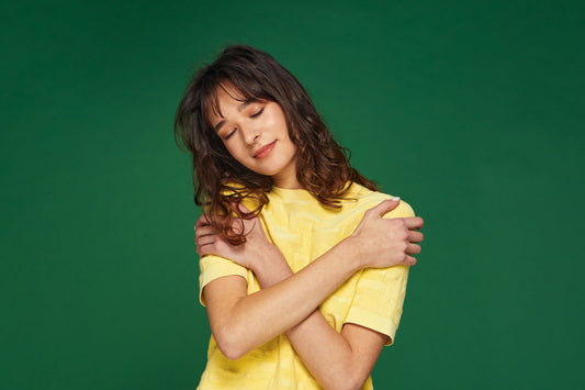 Frau im gelben Shirt vor grünem Hintergund, die sich selbst umarmt