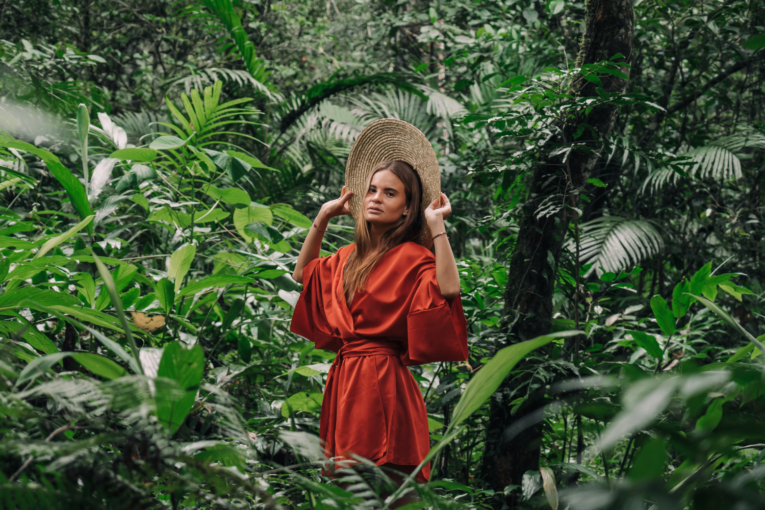 Frau in rotem Kimono aus Baumwollsatin im Dschungel hält ihren Hut hinter ihrem Kopf